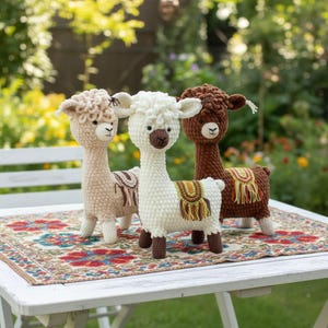 May include: Three crocheted alpaca plush toys standing in a row. The left alpaca is light beige, the center one is white with a dark brown muzzle, and the right one is dark brown. Each features a small, colorful woven saddle with fringe.