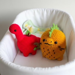 May include: Two handmade crochet toys: a red dinosaur and a yellow creature with green accents. The dinosaur has a small green leaf and brown stem detail. The yellow creature has brown spots and a green spiky crest. Both are in a white crib.