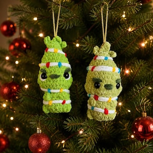 May include: Two handmade crochet ornaments shaped like green creatures with grumpy expressions, black eyes, and dark green eyebrows. Each figure is wrapped in a white yarn strand decorated with small, colourful pom-pom Christmas lights.