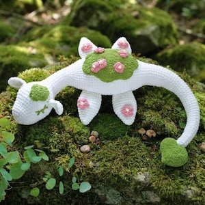 Könnte beinhalten: Ein weißes, gehäkeltes Plüschtier in Form eines langhalsigen Wesens mit einem grünen Fleck auf dem Rücken und dem Kopf. Das Spielzeug hat kleine, gehäkelte rosa Blumen auf den grünen Flächen und Flossen sowie grüne, aufgestickte Augen.