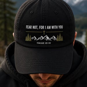 May include: A black baseball cap featuring white embroidered text that reads FEAR NOT, FOR I AM WITH YOU, a small cross symbol, a graphic of white mountain peaks, green pine trees, and the text ISAIAH 41:10 centered on the front panel.