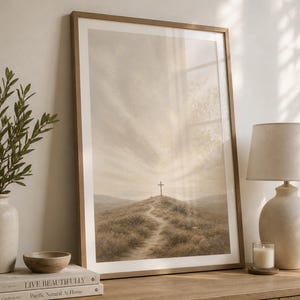 May include: A framed vertical wall art print featuring a landscape illustration of a cross standing atop a grassy hill under a cloudy sky. The artwork uses a muted, neutral colour palette of beige, tan, and soft brown tones, encased in a light wood frame.