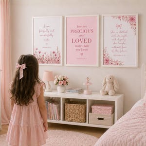 May include: A pink bedroom scene with three framed prints featuring inspirational quotes and floral designs. A young girl in a pink dress stands in front of the prints. The room includes a white shelf, a bed, and decorative items.