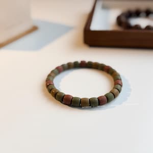 May include: A bracelet made of square beads in shades of green, brown, and red. The bracelet is displayed on a white surface. The words "BEST DIRECTOR" are printed on the bottom right of the image.