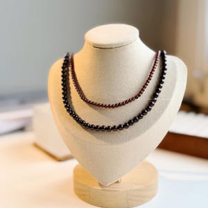 May include: Two beaded necklaces on a mannequin. The top necklace has small, dark red beads, and the bottom one has larger, black beads. The necklaces are displayed on a beige mannequin with a wooden base, showcasing jewellery.