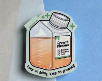 Könnte beinhalten: Ein Aufkleber mit einer Cartoon-Illustration einer durchsichtigen Glasflasche mit einer hellorangen Flüssigkeit darin. Die Flasche ist mit "Growth Medium" beschriftet, darunter stehen die Worte "Ambition, Discipline, Focus". Der Aufkleber hat einen hellgrünen Hintergrund und den Text "Keep on going, keep on growing!" unten.
