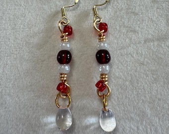 May include: A pair of dangle earrings featuring gold-tone wire hooks and accents. Each earring is composed of a vertical arrangement of small red and white beads, a larger dark red bead, and a clear, teardrop-shaped glass charm at the base.