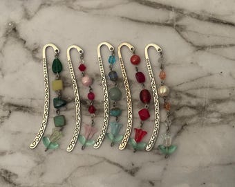 May include: A collection of six beaded bookmarks with silver-toned metal accents. Each bookmark features a different color scheme of beads, including green, pink, blue, and red. The bottom of each bookmark has a small, clear, flower-shaped charm.