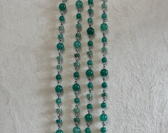 May include: Two long necklaces featuring a series of round and square green beads in varying shades of emerald and mint. The beads are linked together by thin silver-toned metal wire, creating a delicate chain design with a simple hook clasp at the top.