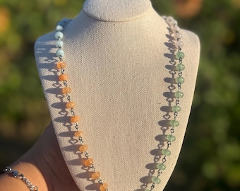 May include: A necklace featuring a central iridescent baroque pearl flanked by a strand of orange beads on one side and a strand of light green beads on the other, all connected by a silver-toned metal chain with a lobster claw clasp closure.