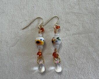 May include: A pair of dangle earrings featuring gold-tone wire hooks, white ceramic beads with yellow sunflower patterns, small translucent orange and clear glass beads, and clear teardrop-shaped glass pendants at the bottom of each earring.