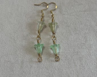 May include: A pair of gold-toned dangle earrings featuring two stacked, bell-shaped glass beads in light green and translucent pale green hues. The beads are connected by gold-toned wire loops and suspended from gold-toned ear hooks.