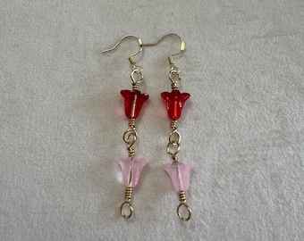 May include: A pair of dangle earrings featuring gold-tone wire hooks and connectors. Each earring displays a translucent red flower-shaped bead positioned above a translucent light pink flower-shaped bead, connected by gold-tone metal wire loops.