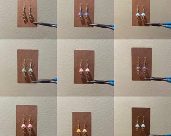 May include: A collection of handmade mushroom earrings in various colors, including green, pink, blue, and yellow. Each pair is displayed on a small brown card. The earrings have a delicate design with gold-colored hooks.