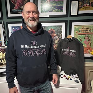 May include: A man wearing a navy blue hoodie with the text "THE SPACE BETWEEN PODCAST" and a graphic of people with headphones. A second gray hoodie with the same design is on a hanger. The background features framed posters and a white cabinet with guitar picks.