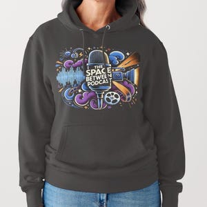May include: Dark gray hoodie with a colorful graphic design. The design features a microphone, film reel, and the text "THE SPACE BETWEEN PODCAST" in white and blue. The hoodie has a front pocket and a hood.