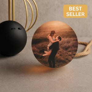 Può includere: Una foto rotonda e illuminata con una coppia che si abbraccia, sullo sfondo di un caldo tramonto. L'immagine è etichettata "BEST SELLER" ed è attaccata a un cordino decorativo. Sullo sfondo c'è un oggetto sferico nero con fori per altoparlanti.