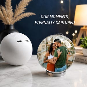 Op de afbeelding: Een wit bolvormig apparaat met twee poorten staat naast een ronde glazen standaard met een foto van een omhelzend koppel. De tekst "OUR MOMENTS, ETERNALLY CAPTURED" is erboven zichtbaar. De objecten staan op een wit oppervlak.