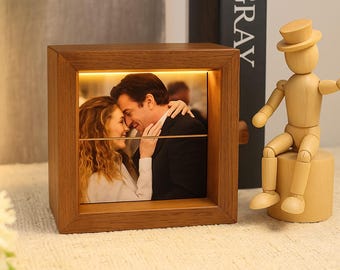 May include: A wooden photo frame with a warm, glowing light. The frame displays a photo of a couple embracing. A wooden artist's mannequin sits on a small wooden cylinder next to the frame. The word "GRAY" is visible on a book in the background.