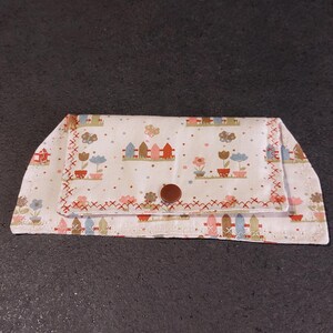 Può includere: Un portafoglio in tessuto bianco con un motivo ripetuto di recinzioni, fiori e case in colori pastello. Il portafoglio ha una chiusura con bottone marrone e cuciture rosse sui bordi.