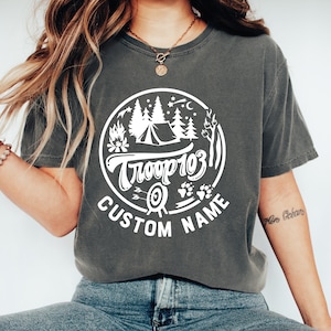 May include: A gray t-shirt with a white graphic design of a campfire, tent, trees, and stars in a circle. The text "Troop 103" is inside the circle, and the text "CUSTOM NAME" is below the circle.