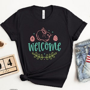 Pu&ograve; includere: Maglietta nera con un coniglio rosa, due uova di Pasqua rosa e il testo "WELCOME" in verde turchese con un design di ramo di foglie verdi.