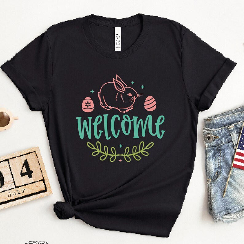Pu&ograve; includere: Maglietta nera con un coniglio rosa, due uova di Pasqua rosa e il testo "WELCOME" in verde turchese con un design di ramo di foglie verdi.