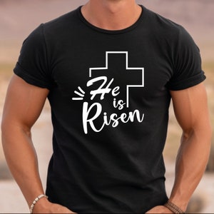 Może przedstawiać: Czarny T-shirt z białym krzyżem i napisem "He is Risen" w białym, ozdobnym pismie.