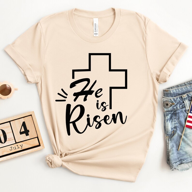 Może przedstawiać: Beżowa koszulka z czarnym krzyżem i napisem "He is Risen" napisanym kursywą.