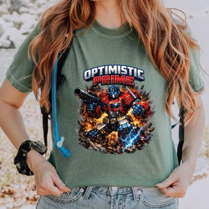 May include: Sage green t-shirt featuring a graphic of Optimus Prime with the text "Optimistic Prime." The robot is depicted in red, blue, and silver, holding a weapon, with a fiery background and lightning bolts.