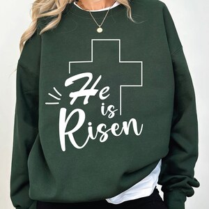 May include: A dark green sweatshirt with a white cross and the text "He is Risen" printed on the front.