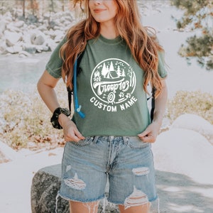 May include: A green t-shirt with a white circle graphic featuring a campfire, tent, trees, and the text "Troop 103" and "CUSTOM NAME".