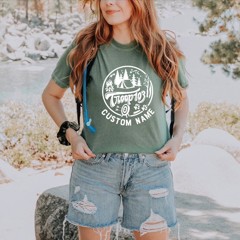 May include: A green t-shirt with a white circle graphic featuring a campfire, tent, trees, and the text "Troop 103" and "CUSTOM NAME".