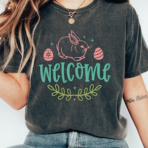 Pu&ograve; includere: Una maglietta grigia scura con un coniglio rosa, due uova rosa e foglie verdi. Il testo "welcome" &egrave; scritto in turchese.