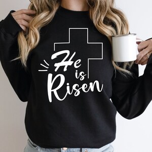 May include: Black sweatshirt with a white graphic design of a cross and the text "He is Risen".