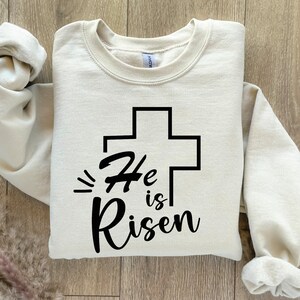 May include: A beige sweatshirt with a black cross and the text "He is Risen" printed on the front.