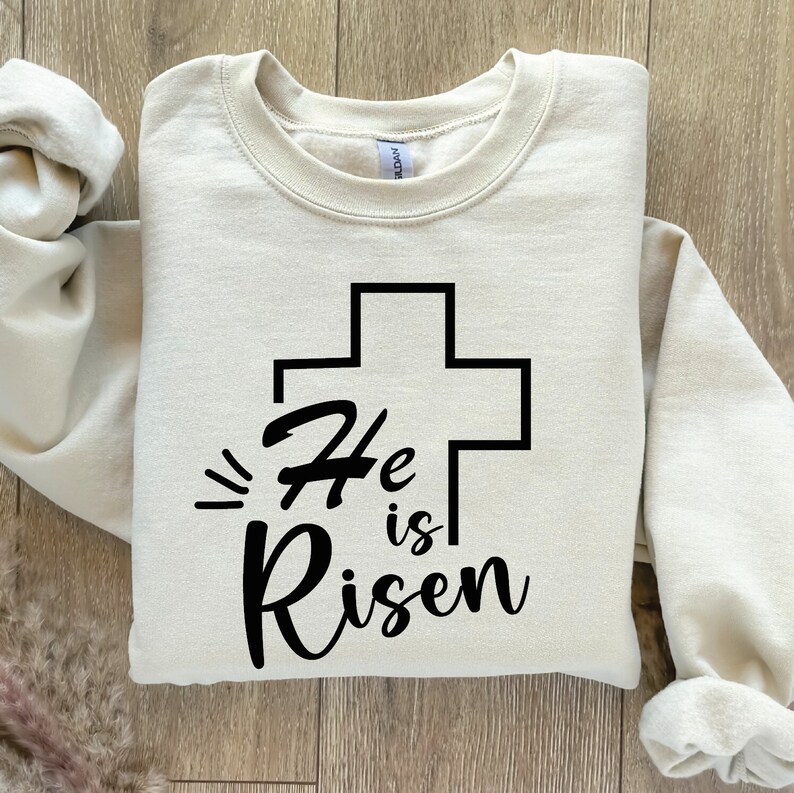 May include: A beige sweatshirt with a black cross and the text "He is Risen" printed on the front.