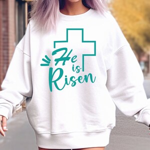 May include: White sweatshirt with a teal green cross and the text "He is Risen" printed on the front.
