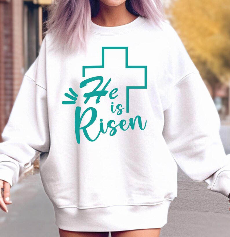 May include: White sweatshirt with a teal green cross and the text "He is Risen" printed on the front.