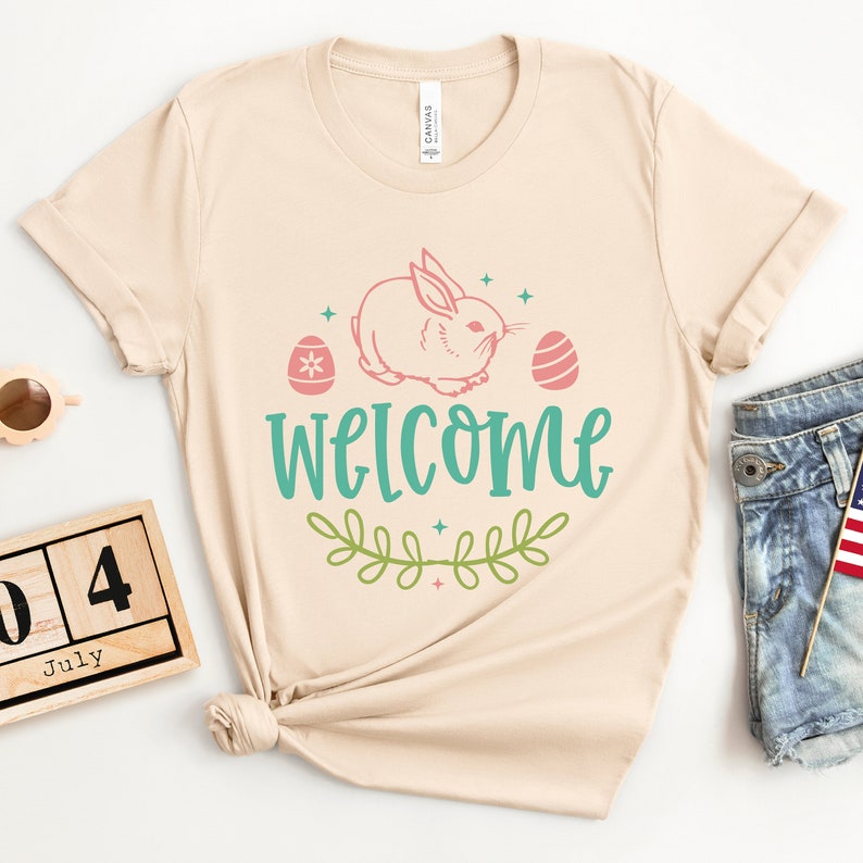 Pu&ograve; includere: Una maglietta beige con un coniglietto rosa, foglie verdi e il testo "WELCOME" in blu turchese. Due uova di Pasqua rosa e bianche sono su entrambi i lati del coniglietto.