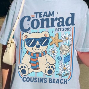 Puede incluir: Camiseta azul claro con un oso con gafas de sol y bufanda, rodeado de estrellas de mar, conchas y flores. El texto dice "Team Conrad, Est 2009, Cousins Beach". La camiseta tiene un corte holgado.