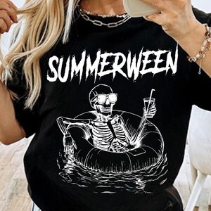 May include: Black t-shirt with the word "SUMMERWEEN" in white, above a skeleton in sunglasses relaxing in a pool float with a drink. The design is a fun, spooky summer theme.