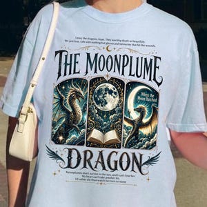 May include: Pale blue t-shirt with a graphic design featuring a dragon, moon, and the text "THE MOONPLUME DRAGON." The design includes illustrations and text. The shirt has short sleeves and a relaxed fit.