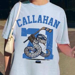 May include: Light blue t-shirt with the word "Callahan" in blue block letters. The shirt features a cartoon snake wearing a hockey helmet and holding a hockey stick, with the number "71" and a hockey puck.