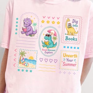 May include: Light pink t-shirt featuring colorful dinosaur illustrations reading books, with text that reads "Dig into Books" and "Unearth Your Summer." The shirt has a relaxed fit and a playful, summery theme.