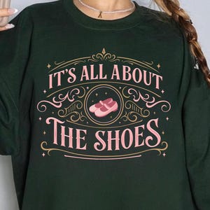 Peut inclure: Sweat-shirt vert foncé avec l'inscription "IT'S ALL ABOUT THE SHOES" en rose et or. Le motif comprend une illustration de chaussures roses. Les mots "HEART BRAVE ART" sont imprimés en bas.