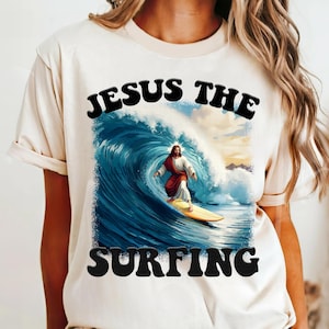 May include: A white t-shirt with the text "JESUS THE SURFING" in black letters. The graphic features a depiction of Jesus surfing on a wave.