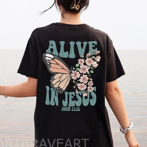 Può includere: Maglietta nera con un design a farfalla e fiori. Il testo "Alive in Jesus" e "John 11:25" è stampato sulla maglietta.