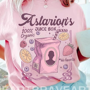 May include: Pale pink t-shirt featuring a juice box graphic with the text "Astarion's 100% Organic Juice Box." The design includes fruit illustrations and the words "his favourite." The shirt also has the text "HEARTBRAVEART" at the bottom.