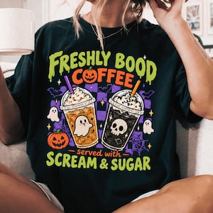 May include: A black t-shirt featuring a graphic print with the text "FRESHLY BOOD COFFEE" and "SERVED WITH SCREAM & SUGAR". The design includes two iced coffee cups, one with a ghost and one with a skull, surrounded by pumpkins, bats, and webs.